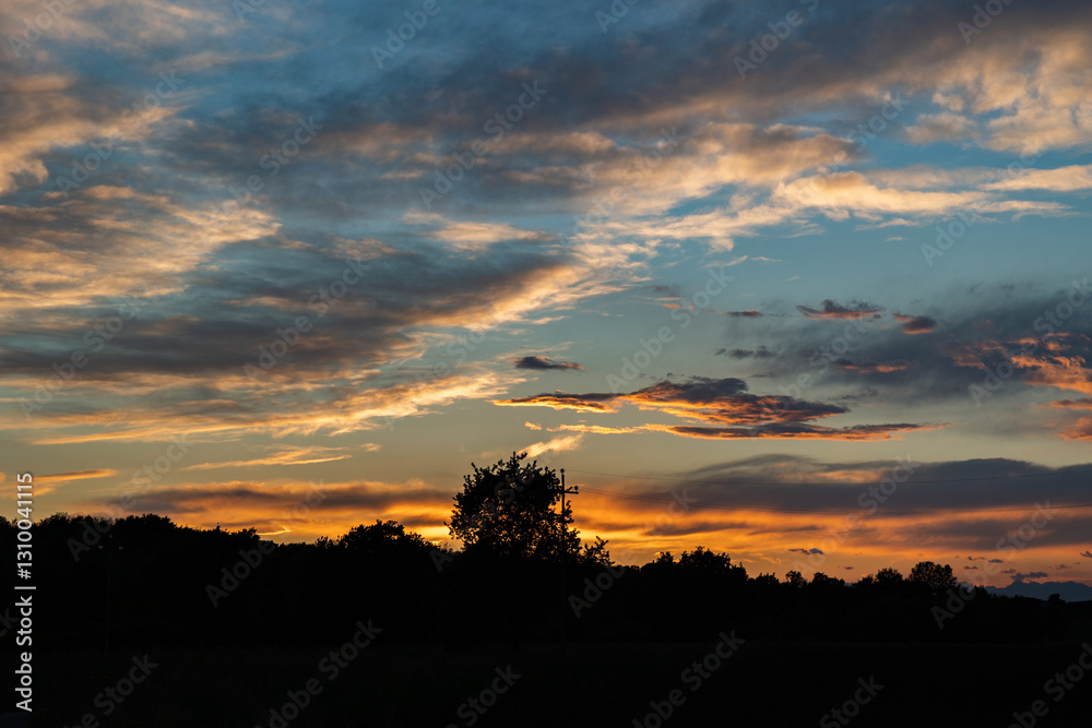 sagome nere di colline ed alberi in primo piano con cielo nuvoloso al tramonto, colorato di arancione
