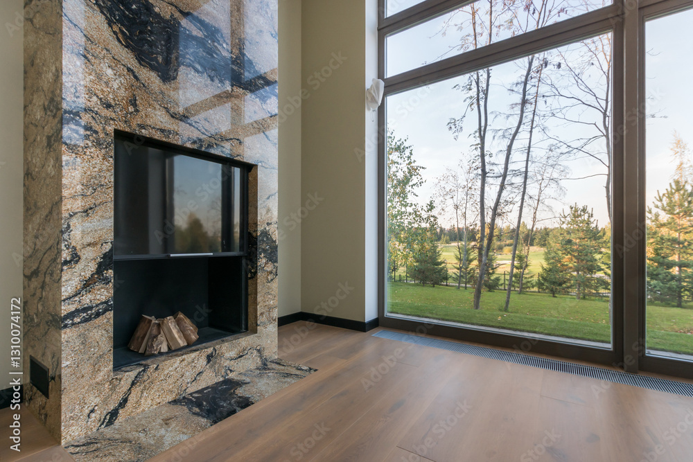 Fototapeta premium Fireplace decorated with marble tiles in the interior of the living room of a private house. Large panoramic window with a view of trees and a green lawn.