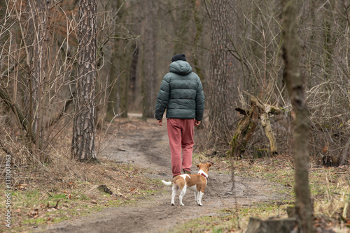 A person wearing a green jacket and red pants strolls down a dirt trail in a forest, accompanied by a small dog. The atmosphere is serene and tranquil, with bare trees around
