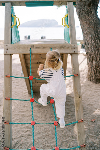 Little girl climbs a rope ladder onto a slide on a beach playground. Back view