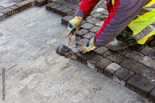 Worker Installing Cobblestone Pavement with Rubber Mallet. Construction worker in safety gear laying cobblestone pavement using a rubber mallet on a sand base.