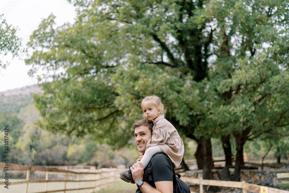 Obraz premium Smiling dad looks back while walking with a little girl on his shoulders through a green garden