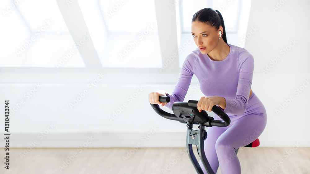 Focused sporty lady training on stationary bike at home and listening music in earbuds, having domestic workout, panorama with copy space, banner Focused sporty lady training on stationary bike at home and listening music in earbuds, having domestic workout, panorama with copy space, banner