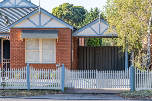 An Australian suburban home with red brick exterior walls, trim on the roof gables, and window awnings. A picket fence surrounds the front yard, with a single carport attached. A modest dwelling.