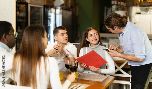 During communicating with girl customer, man waiter writes down order of dishes and drinks, answers customers questions about offer from chef. Employee serve couple clients in cafe