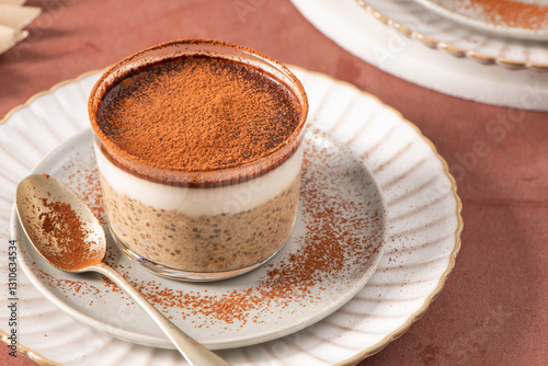 Tiramisu chia pudding in a glass, topped with cocoa powder, served on a white plate with a spoon, creating a visually appealing dessert presentation