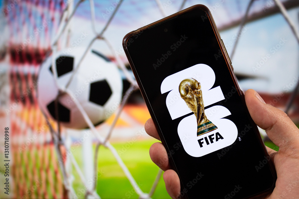 Fototapeta premium A male hand holding a smartphone with fifa world cup 2026 logo over a black background and a football goal blurred background