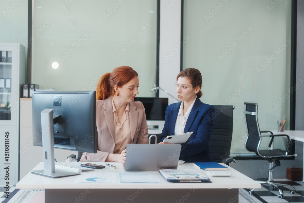 Two Caucasian women work in an office, happily using laptops and computers at a desk, discussing financial projects, analyzing financial statements,collaborating in a meeting to ensure effective