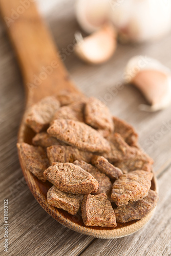Dehydrated chunks of soy meat or texturized vegetable protein on wooden spoon, garlic cloves in the back (Selective Focus, Focus one third into the spoon) 