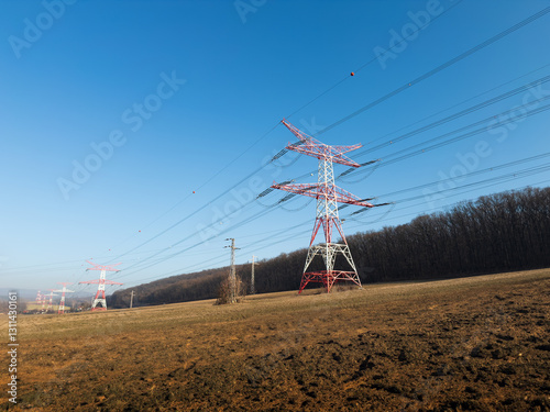 A landscape with high voltage power lines supported by large red and white transmission towers. The towers stretch across a field with dry soil, a wooded area and a clear blue sky.