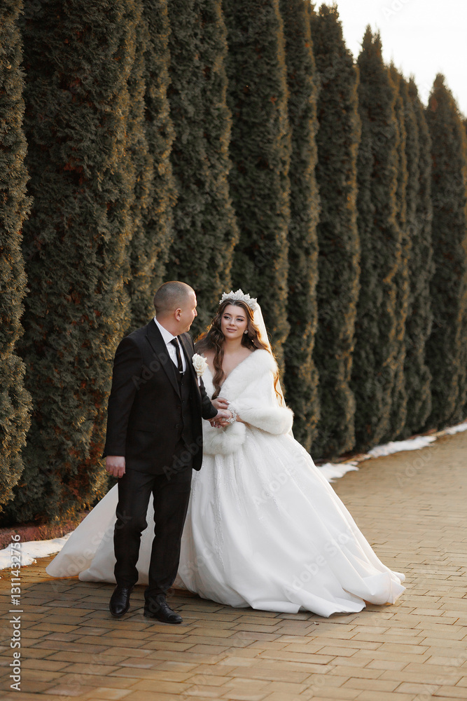 A bride and groom are standing in front of a hedge. The bride is wearing a white dress and a white fur stole A bride and groom are standing in front of a hedge. The bride is wearing a white dress and a white fur stole
