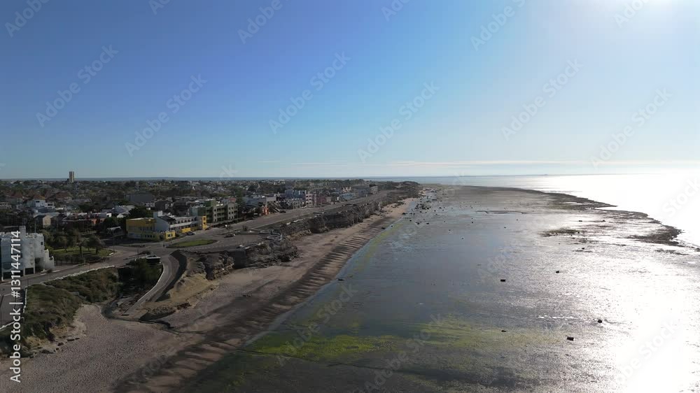 An aerial view of Las Grutas beach, showing the coastline with its distinct rocky formations and sandy beach. The road runs parallel to the shore, surrounded by buildings and residential areas. An aerial view of Las Grutas beach, showing the coastline with its distinct rocky formations and sandy beach. The road runs parallel to the shore, surrounded by buildings and residential areas.