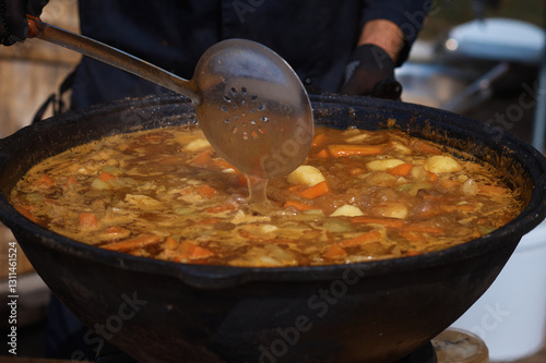 The cook stirs a thick, delicious soup in a large cast iron cauldron with a slotted spoon. National Asian food. Cooking meat dishes outdoors. Selective focus.