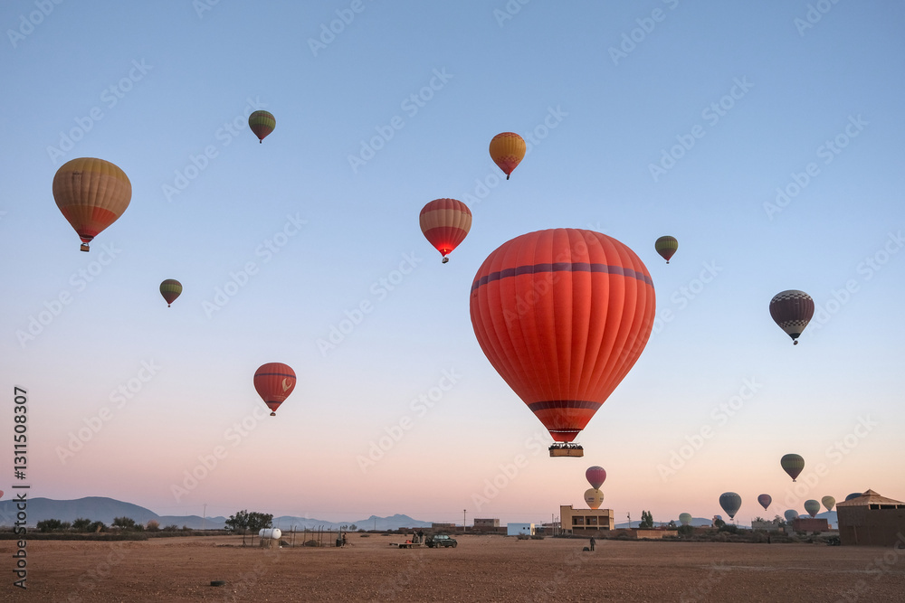 Fototapeta premium Hot air balloons float above an open landscape at sunrise, with mountains in the background. The sky transitions from deep blue to warm hues of dawn.