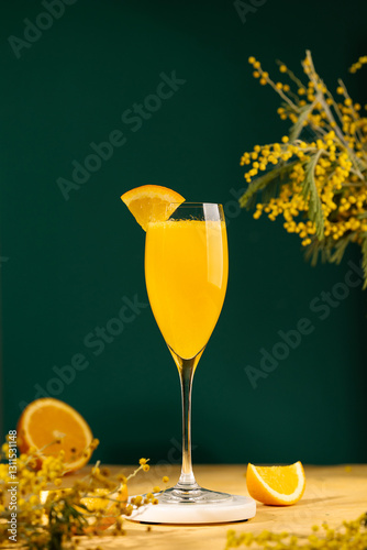 Mimosa cocktail in a champagne glass garnished with a piece of orange. Alcoholic drink made from fresh orange juice and sparkling wine. Yellow table, dark green background. Copy space 