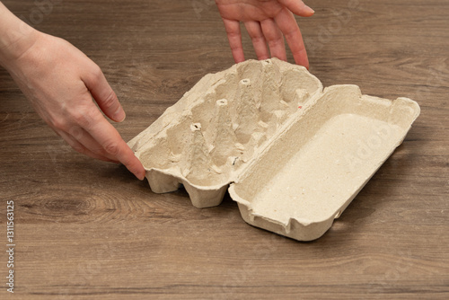 Two hands are positioned near an empty egg carton resting on a wooden surface, showcasing its simple design. The warm light suggests a daytime setting in a kitchen