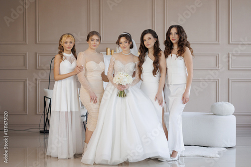 A group of five women are posing for a picture in a room with a white wall. One of the women is wearing a white dress and holding a bouquet of flowers. The other women are also dressed in white