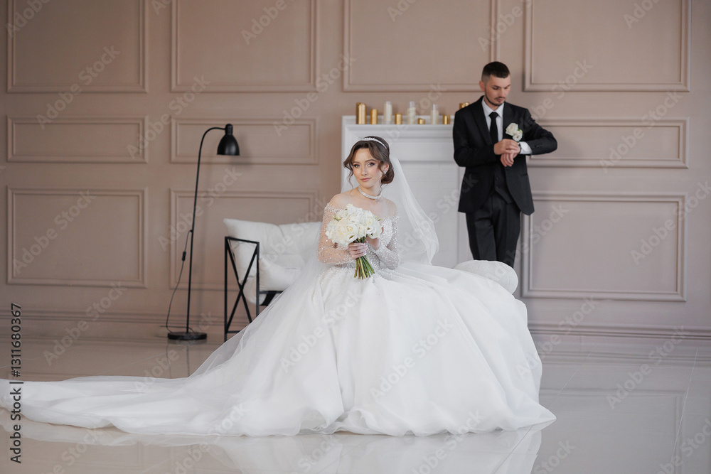 Naklejka premium A bride and groom are posing for a picture in a room with white walls. The bride is wearing a white dress and holding a bouquet of flowers. The groom is wearing a suit and tie