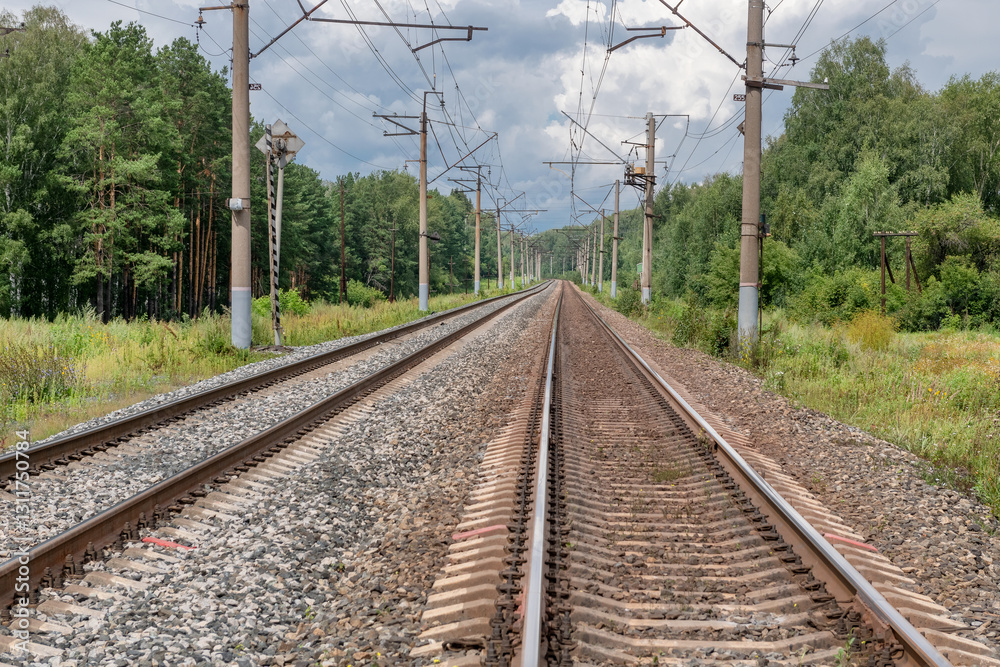 Straight railway tracks cutting through the forest and going off into the distance under a cloudy sky. Overhead power lines create a sense of symmetry and travel through nature. Straight railway tracks cutting through the forest and going off into the distance under a cloudy sky. Overhead power lines create a sense of symmetry and travel through nature.