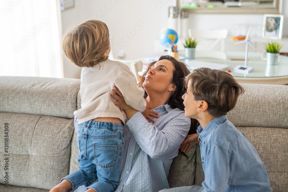 Obraz premium Mother is holding her youngest son, playing with him on the sofa, while her older son is sitting next to them, enjoying the family moment in their living room