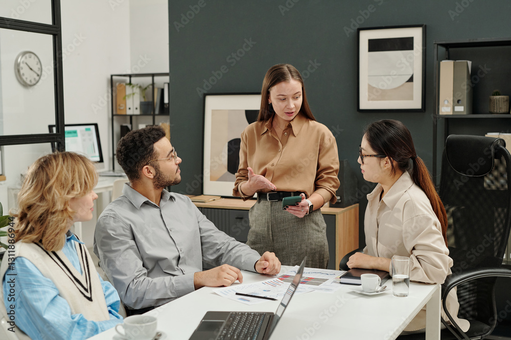 Fototapeta premium Portrait of young businesswoman as female boss pointing at smartphone screen while discussing marketing in business meeting with team in office 