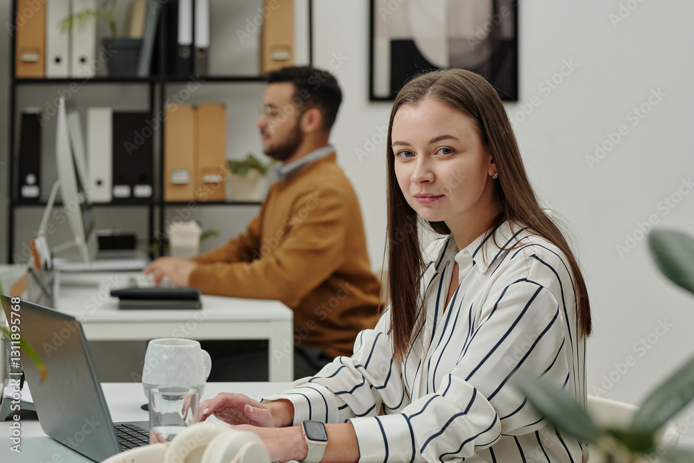 Naklejka premium Portrait of of young woman looking at camera while sitting at workplace in modern office and using laptop copy space 
