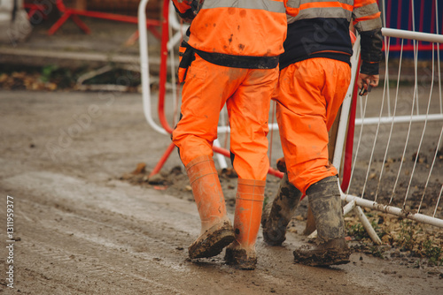 Builders in bright orange uniforms walking on  muddy construction site 