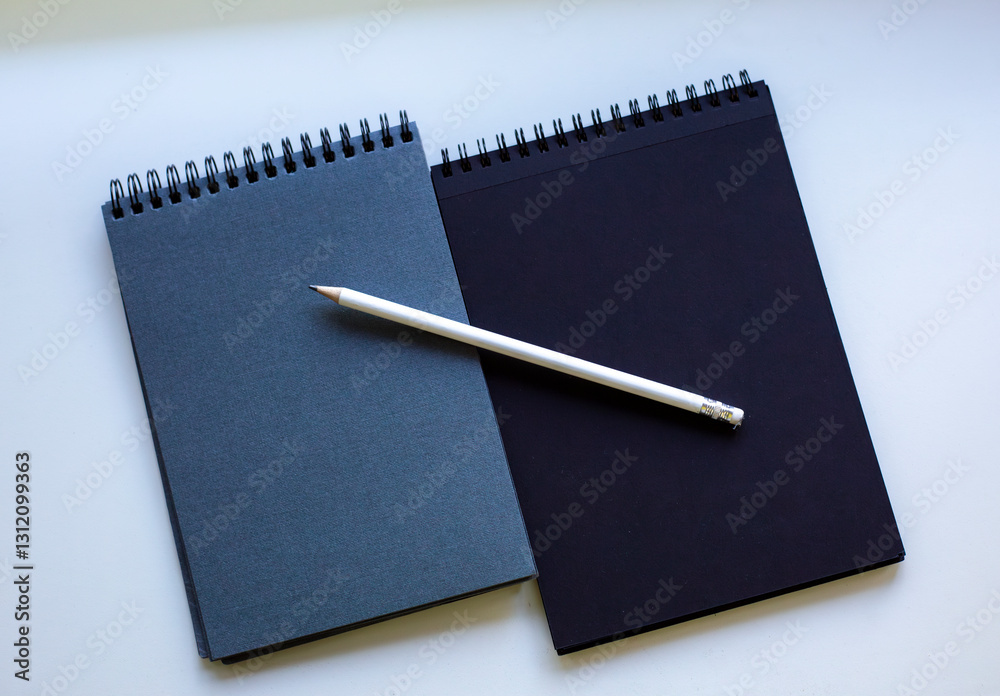 Two spiral-bound notebooks, one dark blue and one black, with a white pencil placed diagonally on them, resting on a white surface. Concept of writing Two spiral-bound notebooks, one dark blue and one black, with a white pencil placed diagonally on them, resting on a white surface. Concept of writing
