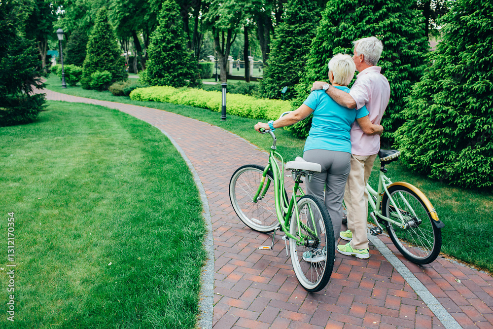 Fototapeta premium A senior couple takes a break on their bicycles, embracing in a peaceful park surrounded by greenery and walking paths