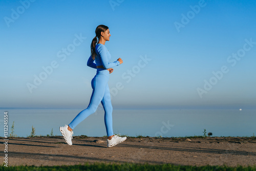 A woman in a vibrant blue outfit briskly walks along a serene waterside path, basking in the calmness of a clear morning sky.