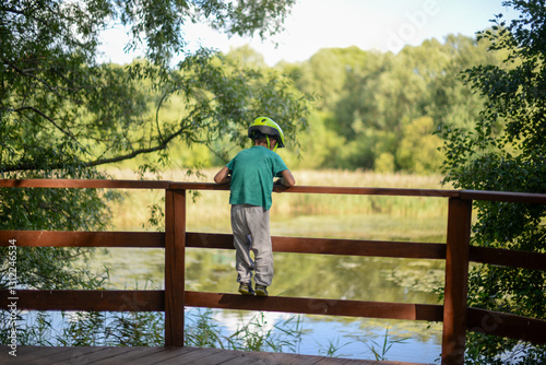 Boy in helmet bravely climbs fence, showcasing spirit of adventure and youthful energy. Vibrant moment of outdoor activity and fun in park nearby the lake in summer sunny day.
