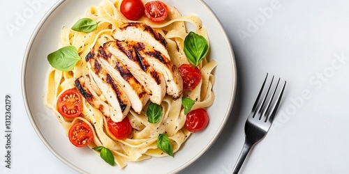Overhead view of grilled chicken breast sliced atop fettuccine pasta with cherry tomatoes and basil, on a white plate, next to a fork