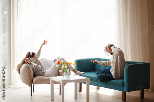 Two  teenagers girls  is relaxing at home in the living room in headphones, sitting in an armchair and sofa,  listening music in headphones