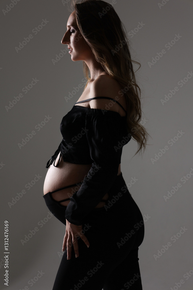 Fototapeta premium Portrait of attractive pregnant woman in black costume, trousers, top, studio photoshoot. Fashion photo of pregnancy. Young pretty girl with long hair touching her belly. Isolated, grey background
