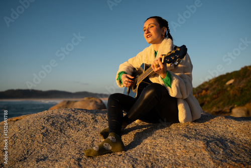 Female musician playing acoustic guitar, perched on rocky coastline during golden sunset, creating melodic sounds against scenic maritime background