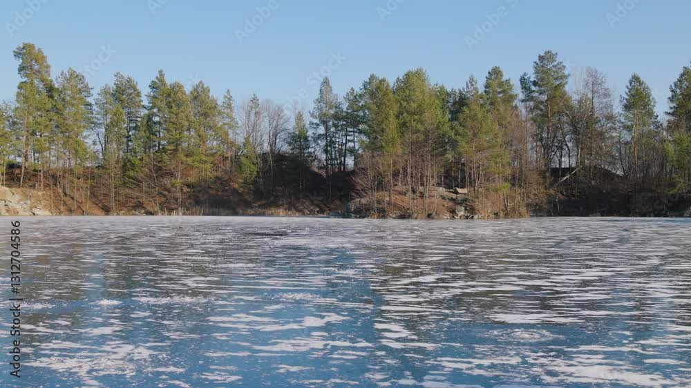 A frozen lake with a textured icy surface reflects the clear blue sky. Surrounding it, tall evergreen and leafless trees stand against a rocky shoreline, creating a serene winter landscape. A frozen lake with a textured icy surface reflects the clear blue sky. Surrounding it, tall evergreen and leafless trees stand against a rocky shoreline, creating a serene winter landscape.