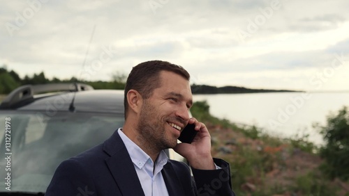 A business man stands near his car on the coast of a lake, sea or ocean, speaks on the phone, he is happy to new successes in his business photo