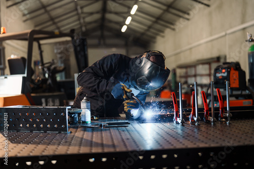 Welder wearing protective gear is using a welding torch to join metal pieces on a welding table in a factory, creating sparks and bright light, demonstrating industrial manufacturing processes