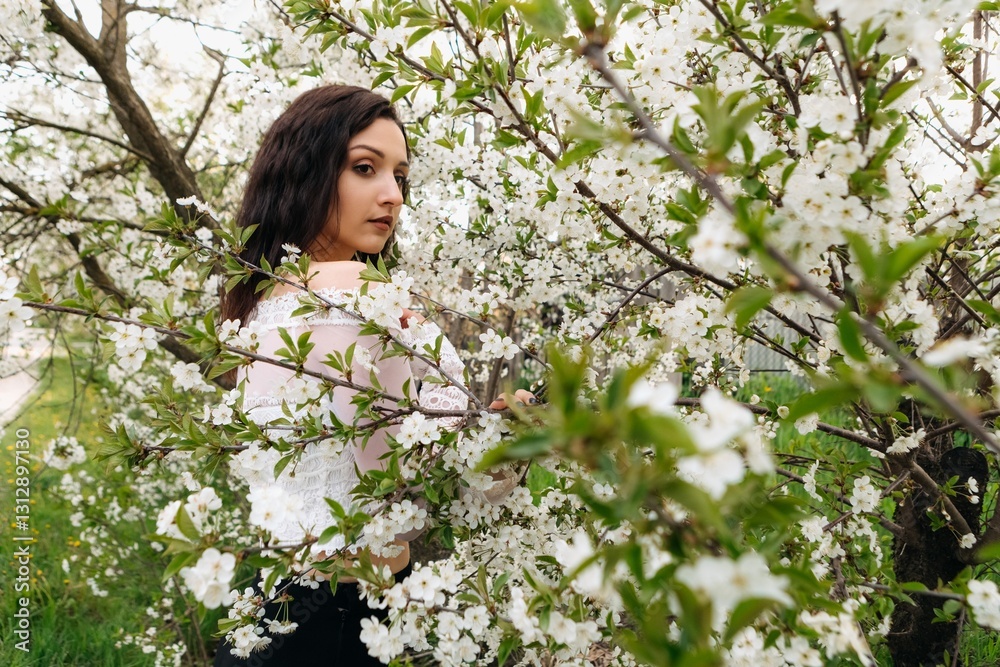 Young woman with long brown hair, wearing a white top, stands amidst a blooming cherry tree, gently touching the delicate white flowers and enjoying the beauty of the spring season Young woman with long brown hair, wearing a white top, stands amidst a blooming cherry tree, gently touching the delicate white flowers and enjoying the beauty of the spring season