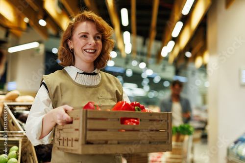 Fotografie Smiling greengrocer carrying wooden crate full of red bell peppers in supermarke