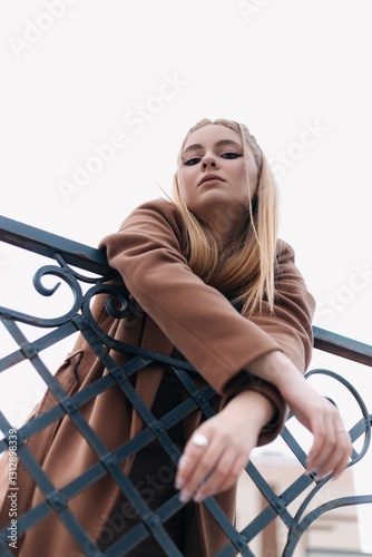Low angle view capturing a fashion model leaning confidently against a railing, showcasing a stylish brown coat that perfectly complements her chic and elegant look