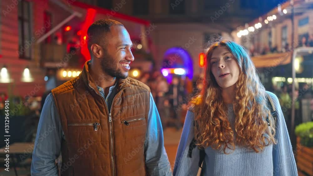 Camera view of loving mixed-raced couple walking down of night city. Handsome man holding hand of his girlfriend during romantic date. People joyfully smiling and communicating with each other. Camera view of loving mixed-raced couple walking down of night city. Handsome man holding hand of his girlfriend during romantic date. People joyfully smiling and communicating with each other.