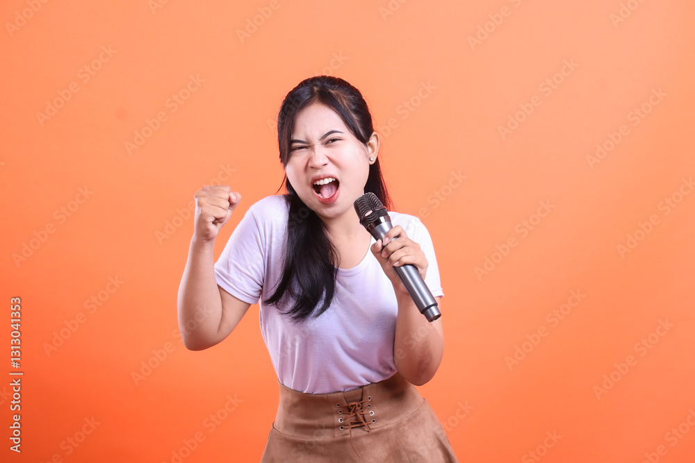An energetic young woman sings passionately into a microphone, raising a fist in excitement while performing, isolated on orange background. An energetic young woman sings passionately into a microphone, raising a fist in excitement while performing, isolated on orange background.