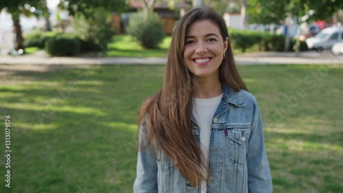A young woman with long brown hair and a radiant smile stands outdoors in a green park. She is wearing a denim jacket over a white sweater and gently touching her hair while looking at the camera