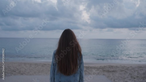 A young woman with long brown hair stands on the beach, gazing at the vast ocean under a cloudy sky. The serene and contemplative scene evokes a sense of peace, solitude, and reflection