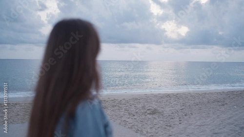 A girl with long brown hair stands on the beach, gazing at the vast ocean under a cloudy sky. The serene and contemplative scene evokes a sense of peace, solitude, and reflection
