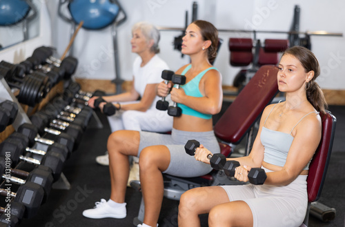 Young woman during grueling workout with dumb-bell in sports hall of fitness club. Intense exercise, increased endurance. Group and personal training, development of individual couching programs