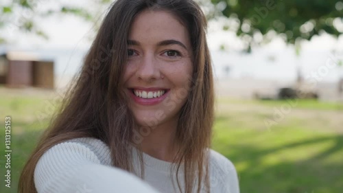 A young woman with long brown hair smiles warmly while standing in a park. She enjoys the fresh air and natural surroundings, radiating happiness and relaxation.