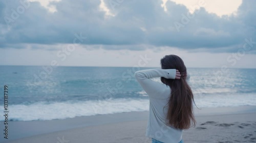 A young woman with long brown hair stands on the beach, facing the ocean with her hands in her hair. Dressed in a cozy sweater and jeans, she enjoys the peaceful moment, embracing the fresh sea breeze
