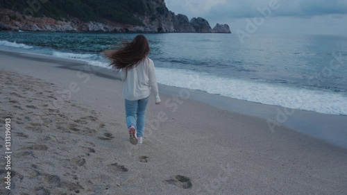 A girl with long brown hair runs barefoot along the sandy beach, enjoying the fresh sea breeze. Dressed in a cozy sweater and jeans, she embraces freedom and joy, with waves crashing gently nearby